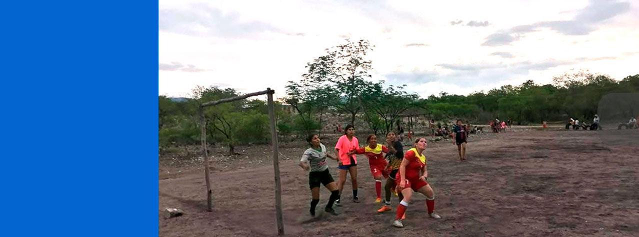 Mujeres jugando un partido de futbol, balón va en el aire frente al arco para poder anotar un gol. En pondores